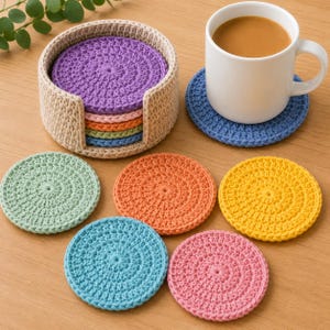 May include: A set of round, crocheted coasters in various colors including purple, pink, orange, green, blue, and yellow. The coasters are stored in a beige, circular, crocheted holder. One blue coaster sits under a white ceramic mug filled with coffee.