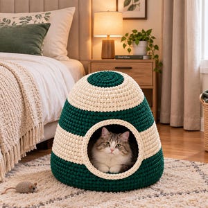 May include: A dome-shaped pet bed made of thick, crocheted yarn in alternating horizontal stripes of dark forest green and cream. The bed features a circular opening at the front, inside of which sits a fluffy, long-haired cat with grey and white fur.