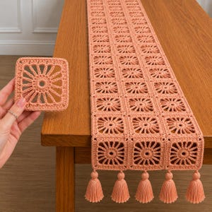 Puede incluir: Un camino de mesa de ganchillo color melocotón hecho de motivos cuadrados repetidos con un patrón central de rayos de sol. El camino presenta cinco borlas a juego en el borde inferior. Se sostiene por separado un motivo cuadrado a juego.