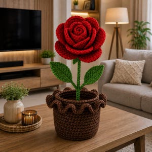 May include: A large, vibrant red crocheted rose flower with a green stem and two green leaves, planted in a dark brown crocheted pot. The pot features a decorative ruffled top edge, and the entire piece is crafted from yarn with visible stitching.