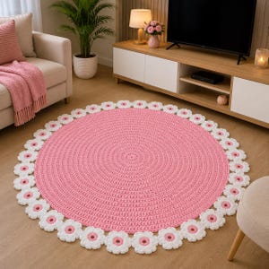 May include: A circular crochet rug featuring a solid pink center with a textured stitch pattern, surrounded by a decorative border of white flower-shaped motifs with pink centers. The rug is crafted from yarn and lies flat on a wooden floor surface.