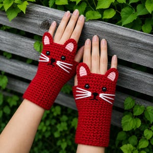 May include: A pair of fingerless gloves crocheted in red yarn, featuring a cat face design with two pointed ears, black button eyes, a small black embroidered nose, and three white yarn whiskers on each side, with light pink inner ears.