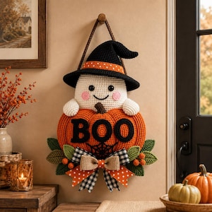 Pode incluir: Uma decoração de parede em crochê apresentando um fantasma branco usando um chapéu de bruxa preto com uma faixa laranja de bolinhas. O fantasma segura uma grande abóbora laranja com o texto preto BOO e uma teia de aranha, com folhas verdes.