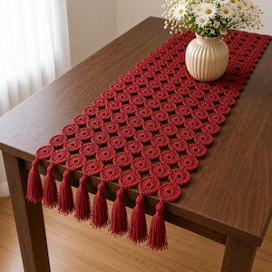 May include: A deep red crochet table runner featuring a repeating circular pattern and finished with matching red tassels along the edge. The runner is constructed from interconnected yarn circles, creating a textured, openwork design that drapes.