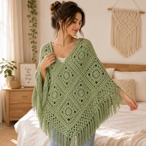 Può includere: Un poncho all'uncinetto color verde salvia con scollo a V e un motivo ripetitivo di quadrati con centri circolari. Il capo è realizzato con quadrati di filato interconnessi ed è rifinito con lunghe frange pendenti lungo il bordo inferiore.