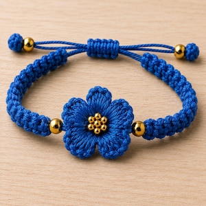 May include: A blue cord bracelet featuring a central five-petaled flower woven from blue thread with a cluster of seven small gold-toned beads at its center. The band includes two additional gold-toned beads and an adjustable sliding knot closure.