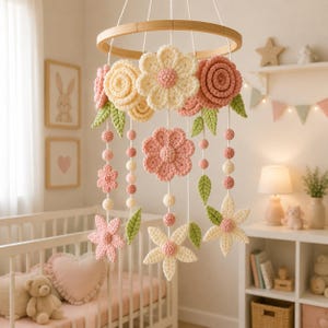 Peut inclure: Un mobile suspendu composé de fleurs et de feuilles crochetées dans des tons crème, rose pâle et vert sauge. Les éléments sont suspendus à un cadre circulaire en bois par des fils blancs, entrecoupés de petites sphères crochetées assorties.