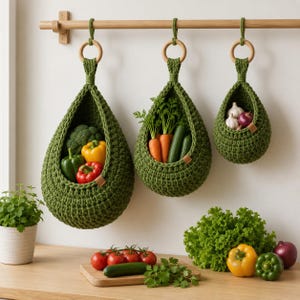 Op de afbeelding: Drie olijfgroene gehaakte hangmanden van verschillende groottes, opgehangen aan een houten stang met houten ringen. De manden bevatten verse producten zoals broccoli, paprika, wortels, komkommers, knoflook en rode uien.