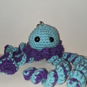 May include: A crocheted octopus keychain featuring a light blue rounded head with two black plastic eyes. The octopus has a ruffled purple collar and multiple spiraling tentacles that alternate between light blue and purple yarn colors.