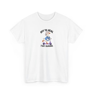 Può includere: Una t-shirt bianca a maniche corte con la grafica di un'oca che indossa una casacca blu, una cuffia chirurgica a motivi e uno stetoscopio mentre guida un monopattino viola. Il testo "OFF TO HEAL" appare sopra e "TINY HUMANS" sotto.