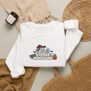 Könnte beinhalten: Ein weißes Langarm-Sweatshirt mit einer Grafik eines Geistes, der einen Hexenhut und eine blaue Gesichtsmaske trägt und medizinische Instrumente hält. Unter dem Geist steht der Text "STERILE PROCESSING" neben einem kleinen orangefarbenen Kürbis.