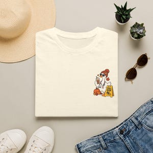 May include: A cream-colored t-shirt featuring a graphic of a goose wearing a beanie and sunglasses, holding a coffee cup and a stethoscope. Next to the goose is a pumpkin and a yellow sign with the text FALL RISK and a falling figure icon.
