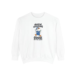 May include: A white long-sleeve sweatshirt featuring a graphic of a goose dressed as a dental assistant wearing a blue scrub top, a patterned head covering, and sunglasses, holding a dental mirror and a tooth, with the text dental assistant GOOSE ON THE LOOSE.