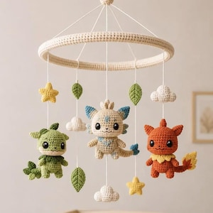 May include: A crochet baby mobile featuring a circular cream-colored frame with hanging yarn figures. Three creatures in green, cream with blue accents, and orange are suspended alongside yellow stars, green leaves, and white clouds on white strings.