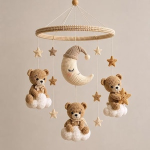 Puede incluir: Un móvil colgante con tres ositos de peluche tejidos a ganchillo sobre nubes blancas, una luna creciente central con cara dormida y gorro beige, y varias estrellas pequeñas tejidas en tonos beige y marrón suspendidas de un aro de madera.