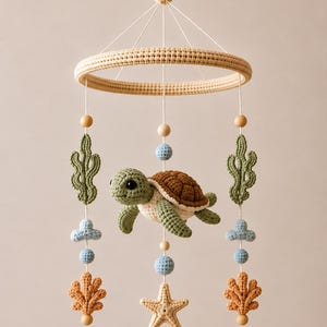May include: A hanging mobile featuring a central crocheted sea turtle with a brown shell and green body. It includes dangling crocheted elements like green seaweed, light blue clouds, tan coral, and a starfish, all suspended from a circular frame.