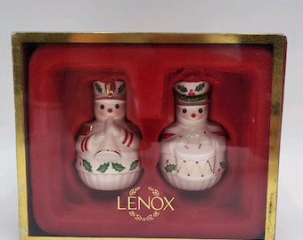 May include: A Lenox boxed set of two porcelain ornaments featuring festive figures in uniform. One figure plays a trumpet, and the other plays a drum. Both are decorated with green holly leaves, red berries, and gold accents, set against a red velvet interior.