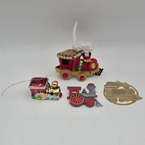 May include: A collection of four train-themed ornaments including a red and yellow felt train with sequins, a small glass locomotive, a flat painted metal train ornament, and a circular gold-toned metal cutout featuring a train silhouette.