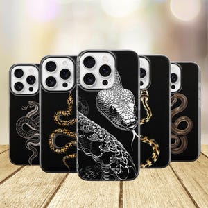 Peut inclure: Cinq coques de smartphone noires alignées, chacune présentant une illustration de serpent différente. Les motifs incluent un dessin au trait blanc, des motifs dorés, une tête de serpent blanche détaillée, un serpent tacheté doré et un serpent brun.