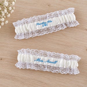 May include: Two white satin wedding garters with delicate white lace trim. The top garter features blue embroidery reading Something Blue S heart J. The bottom garter features blue embroidery reading Mrs. Amell. Both garters are rectangular in shape.
