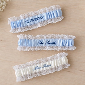 May include: Three elastic garters featuring white lace trim. Two garters are light blue satin with dark blue embroidery reading IXXVIII-MMXXVI and The Smiths. The third garter is white satin with dark blue embroidery reading Mrs. Ross.