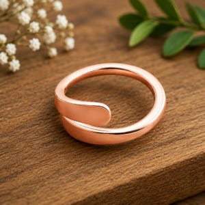 Könnte beinhalten: Ein roségoldfarbener Ring mit glatter, polierter Oberfläche und offenem Bypass-Design. Das Band hat eine abgerundete, durchgehende Form, die sich an der Vorderseite leicht überlappt und für einen klaren, metallischen Glanz sorgt.