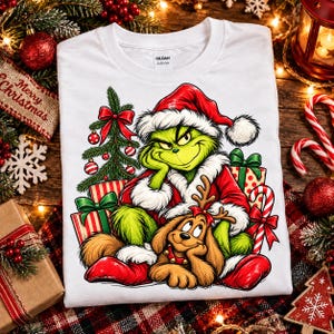 Puede incluir: Una camiseta blanca con el gráfico de un personaje verde con gorro y traje de Papá Noel rojo, sentado junto a un árbol de Navidad decorado y regalos. Un perro marrón con astas de reno está sentado frente al personaje.