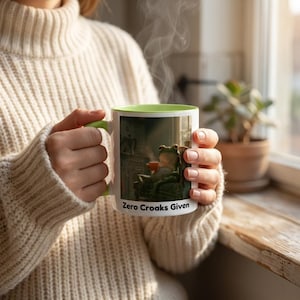 May include: A white ceramic mug with a light green interior and handle, featuring a graphic of a frog sitting in a chair holding a cup. The text "Zero Croaks Given" is printed in black at the bottom of the mug, which is held by two hands.
