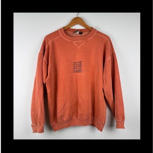 Puede incluir: Una sudadera de manga larga y cuello redondo en color naranja desteñido, con cuello, puños y dobladillo acanalados. El centro del pecho muestra un texto en negro que dice IT'S LIVE. IT'S LIFE. con un texto más pequeño debajo.