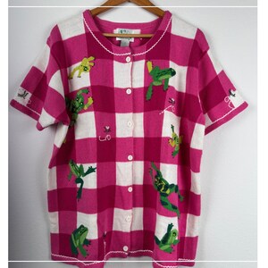 May include: A short-sleeved button-down cardigan featuring a pink and white checkered pattern. The knit garment is decorated with embroidered green frog motifs scattered across the front, finished with a white scalloped trim along the neckline.