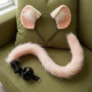 May include: A set consisting of a thin black headband with two large, rounded, pale pink faux fur ears and a long, matching pale pink faux fur tail attached to a black adjustable nylon strap with a plastic buckle and a metal D-ring.
