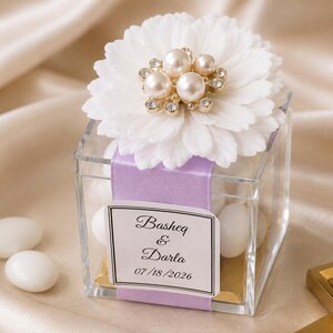 May include: A clear square favour box filled with white almond sweets, wrapped in a lavender ribbon, and topped with a white flower featuring a cluster of faux pearls and clear rhinestones. A label reads Basheq & Darla 07/18/2026.