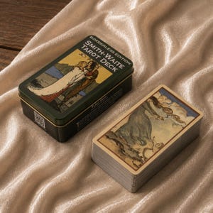 Op de afbeelding: Een rechthoekig donkergroen metalen blik met het label Borderless Edition Smith-Waite Tarot Deck met een illustratie van twee figuren, naast een stapel tarotkaarten met een figuur in een blauwe mantel onder een bewolkte lucht.