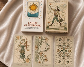 Könnte beinhalten: Ein Radiant Soul Tarot-Kartendeck, ein weißes Tarot-Handbuch und drei Tarotkarten mit den Bezeichnungen The Justice, The Moon und Ten of Cups. Die Kartenschachtel zeigt eine Illustration einer Person mit einer Blume, während das Handbuch ein Sonnensymbol zeigt.