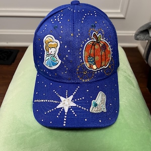 May include: A royal blue baseball cap adorned with scattered clear rhinestones. The front features embroidered patches of a cartoon character, an orange pumpkin carriage, a white starburst, and a single glass slipper, all embellished with crystals.