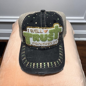 Könnte beinhalten: Eine schwarze, abgenutzte Baseballkappe mit Netzrücken und einem grün-goldenen Aufnäher mit der Aufschrift I WILL TRUST PROVERBS 3:5 sowie einem kleinen Herzsymbol. Das Vorderteil und der Schirm sind mit verstreuten grünen und goldenen Strasssteinen verziert.
