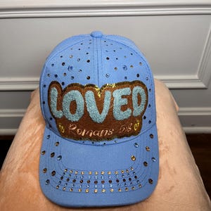May include: A light blue baseball cap featuring a brown patch with the word LOVED in light blue letters and the text Romans 5:8 in gold. The cap is decorated with scattered gold and blue rhinestones across the crown and the curved brim.