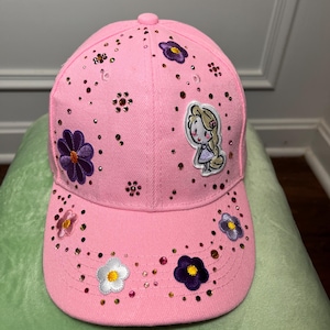 May include: A light pink baseball cap decorated with scattered small rhinestones and several embroidered flower patches in purple, white, and pink. A small embroidered patch of a cartoon character with long blonde hair is positioned on the front.