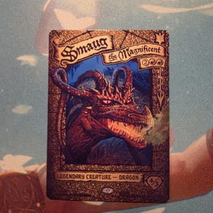 Può includere: Una carta collezionabile rettangolare con l'illustrazione di un drago rosso, il testo Smaug the Magnificent e Legendary Creature - Dragon. La carta presenta un bordo decorativo color oro e mostra un valore di forza e costituzione 4/3.