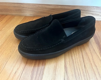 Puede incluir: Un par de mocasines de ante negro con un detalle de costura. Los mocasines tienen una puntera redondeada y una suela plana.