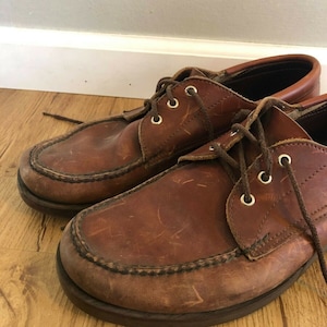 May include: A pair of brown leather lace-up shoes with a moc toe design. The shoes have a worn look and are made of a durable material.
