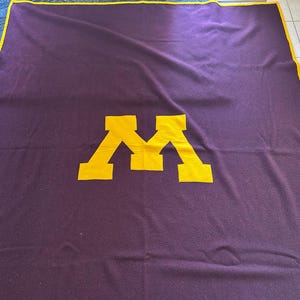 May include: A purple fleece blanket with a large yellow "M" in the center.