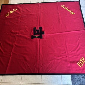 Op de afbeelding: Rode deken met zwarte rand en gele geborduurde tekst. De tekst luidt "Al Reiss Swimming 1972". In het midden bevindt zich een zwart-geel logo.