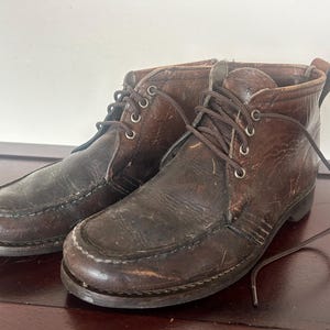 May include: A pair of worn brown leather boots with laces. The boots have a moccasin-style toe and a short ankle height. The leather shows signs of wear, with creases and scuffs. The laces are brown and tied.