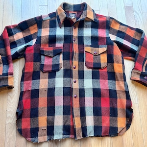 May include: A vintage, button-down plaid shirt in a bold pattern of red, orange, black, and cream. The shirt features two chest pockets, a collar, and buttoned cuffs. The fabric appears to be a thick, warm material, suitable for cooler weather.