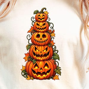 Op de afbeelding: Een crèmekleurig t-shirt met een centrale afbeelding van vijf gestapelde pompoenen. De pompoenen variëren in grootte, hebben elk een uniek uitgesneden gezicht en zijn omringd door groene ranken en oranje en gele herfstbladeren.