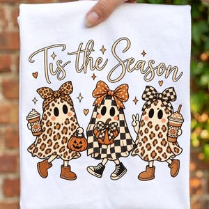 May include: A white t-shirt featuring a graphic of three ghosts wearing bows and boots. The text 'Tis the Season' is printed above them. The ghosts have leopard print and checkered patterns, holding a pumpkin, a jack-o'-lantern, and coffee cups.