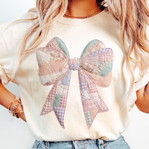 Puede incluir: Una camiseta de manga corta de color crema con un gran gráfico central de un lazo. El lazo presenta un diseño de patchwork en tonos pastel suaves de rosa, azul y lavanda, incorporando estampados florales, de lunares y de cuadros vichy.