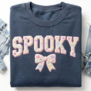 May include: A navy blue crewneck sweatshirt featuring the word SPOOKY in large, cream-colored chenille varsity letters with pink trim. The letters and a central bow graphic are decorated with small, multicolored pastel heart patches.