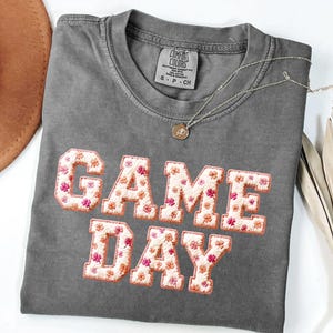 Puede incluir: Una camiseta de manga corta gris carbón con cuello redondo y etiqueta de la marca Comfort Colors. El frente presenta el texto GAME DAY en letras grandes de bloque rellenas con un patrón de tela blanca de pequeñas flores rosas y naranjas.